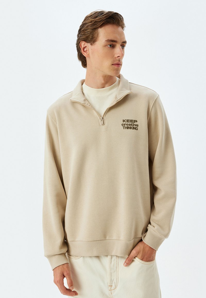 Beige Zip-Up Sweatshirt aus weichem Material mit Rundhalsausschnitt und gerippten Bündchen. Textdruck: „HALTE kreative DENKWEISE.“