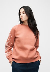 Γυναίκα με μακριά μαύρα μαλλιά που φοράει ροδακινί φούτερ με λευκά λουλουδάτα σχέδια στα μανίκια και σκούρο μπλε παντελόνι, με τα χέρια στις τσέπες, κοιτάζοντας προς τα αριστερά.