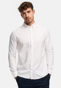 Jeune homme aux cheveux bouclés portant une chemise blanche à boutons et un pantalon sombre, les mains dans les poches, sur un fond clair uni.