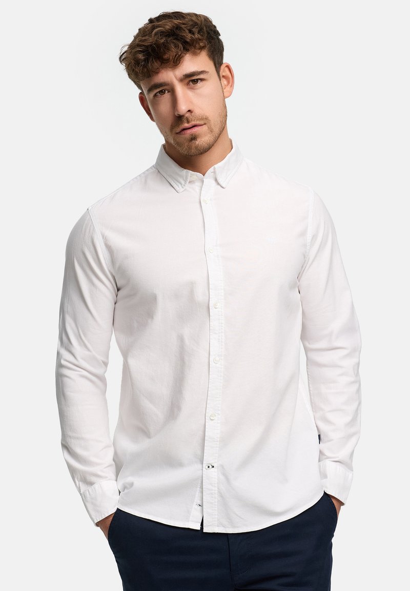 Jeune homme aux cheveux bouclés portant une chemise blanche à boutons et un pantalon sombre, les mains dans les poches, sur un fond clair uni.