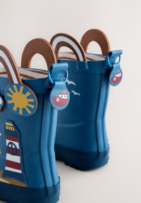 Bottes de pluie bleues pour enfants avec motifs de phare et de soleil, poignées rayées marron et petits tirettes en forme de bateau.