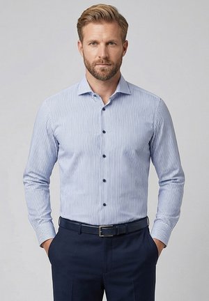 Man met blond haar en baard, in een blauw-wit gestreept overhemd en donkerblauwe broek, staand met handen in de zakken.