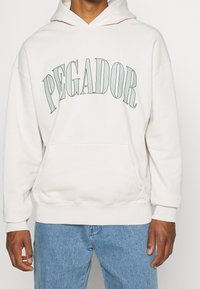 Felpa con cappuccio color crema con la scritta "PEGADOR" in lettere verdi curve, tasca frontale e polsini e orlo a costine. Indossata con jeans blu.