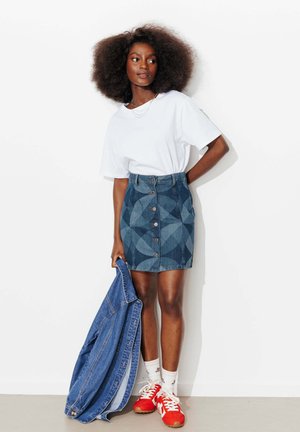 Femme aux cheveux afro portant un t-shirt blanc, une jupe en denim à motifs, des baskets rouges, tenant une veste en denim contre un mur blanc.