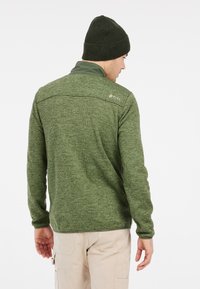 Groene fleecetop met een gestructureerde afwerking, voorzien van een halve rits kraag en een logo-accent op de achterkant. Gecombineerd met een donkergroene beanie.