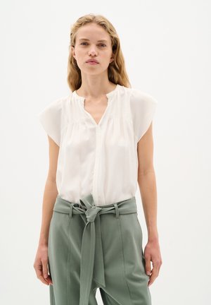 Camicia leggera in crema con maniche corte, scollatura a V e sottili pieghe. Abbinata a pantaloni verdi salvia a vita alta con cintura annodata.