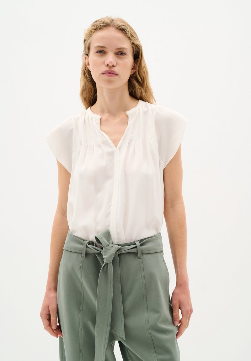 Camicia leggera in crema con maniche corte, scollatura a V e sottili pieghe. Abbinata a pantaloni verdi salvia a vita alta con cintura annodata.