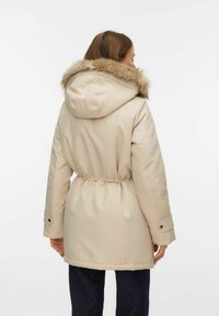 Bēšas krāsas parka ar kažokādas apkakli kapucē, vidukļa saspiešanu un pogām aizdares uz aprocēm. Gluda auduma tekstūra ar vieglu spīdumu.