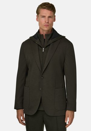 Man met een donker olijfgroene blazer over een hoodie met ritssluiting, staand en recht vooruit kijkend tegen een effen lichte achtergrond.