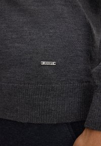 Pull en tricot gris charbon avec un ourlet côtelé et une étiquette en métal argenté "JOOP !" cousue sur le côté. Texture lisse, design classique.