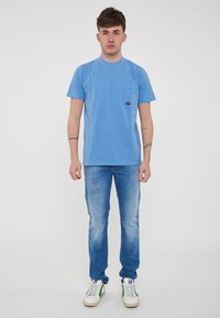 T-shirt azzurra con colletto rotondo e tasca, abbinata a jeans azzurri e sneaker bianche con dettagli verdi.