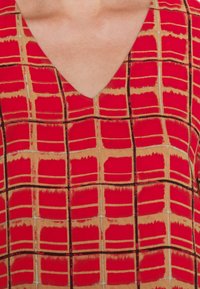 Tela estampada en rojo y beige con un diseño de cuello en V, que presenta una cuadrícula de formas rectangulares contorneadas en negro y suaves acentos en beige.