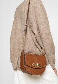 Borsa a tracolla in pelle marrone con una forma curva, hardware dorato, tracolla a catena e finitura texture. Presenta un logo prominente sul davanti.