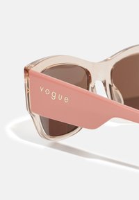 Rosa solbriller med klar ramme, brune linser og gull "vogue"-merkevare på armen. Vinklet design med en glatt tekstur.
