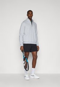 Pullover cinza claro em fleece com meia abertura, calções pretos, segurando uma raquete de tênis e usando ténis brancos com tecido texturizado visível e detalhes azuis.