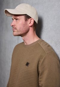 Cappello beige e felpa in tessuto marrone con una patch rotonda sul petto, su uno sfondo grigio.