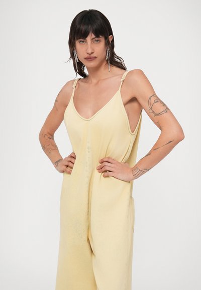 Άτομο με σκούρα μαλλιά και τατουάζ στα χέρια που φοράει χαλαρό αμάνικο ολόσωμο φόρεμα παλ κιτρινού χρώματος, με τα χέρια τοποθετημένα στους γοφούς.