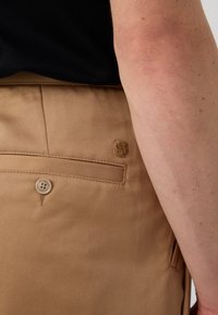 Beige trousers with a smooth texture, featuring a back pocket with a button closure and a subtle embossed logo above it.