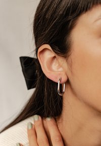 Srebrna kolczyk w kształcie obręczy wiszący z ucha, o minimalistycznym wzornictwie. Włosy ułożone z czarną kokardą, na teksturowanym tle.