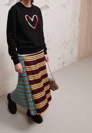 Personne portant un pull noir avec un cœur coloré, une jupe plissée avec des rayures jaunes, bordeaux et turquoise, des bottes noires et tenant un petit sac à motif animalier.