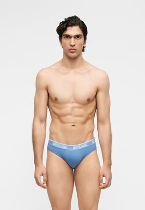 Młody mężczyzna stoi na jednolitym tle, ubrany w niebieskie majtki marki Moschino, patrząc prosto z neutralnym wyrazem twarzy.