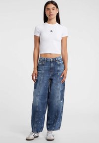 Weißes, kurz geschnittenes T-Shirt mit einem schwarzen Sternlogo, kombiniert mit locker sitzenden, hellblauen, abgenutzten Jeans mit Farbspritzern.