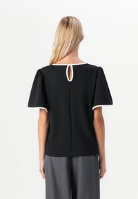 Femme aux longs cheveux blonds portant un haut noir à manches courtes avec bord blanc et une ouverture en forme de clé dans le dos, associé à un pantalon large gris.