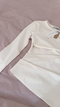 Long-sleeve cream top made of smooth fabric, featuring a fitted design with gathered detail at the waist. Tag attached, laid on a ribbed surface.