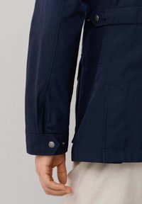 Chaqueta impermeable azul marino con acabado texturizado, que cuenta con cierre de botón a presión en el puño y un ajuste entallado.