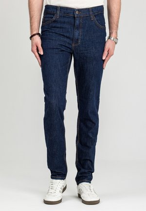 Mężczyzna noszący ciemnoniebieskie dopasowane jeansy, białe trampki, białą koszulę, srebrny zegarek na lewej ręce oraz brązową bransoletkę na prawej ręce.