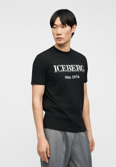 Νεαρός άνδρας με μαύρα μαλλιά, φορώντας μαύρο t-shirt με το κείμενο "ICEBERG since 1974" και γκρι παντελόνι, κοιτάζει προς τα αριστερά του.