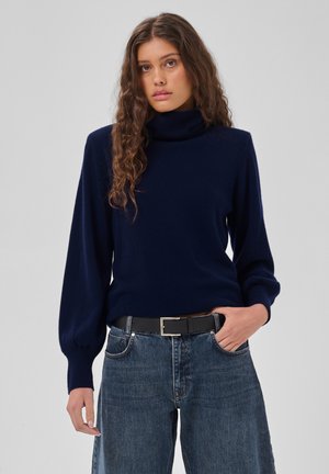Jeune femme aux longs cheveux bouclés portant un pull à col roulé bleu marine et un jean bleu avec une ceinture noire, une main dans la poche.
