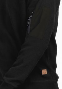 Suéter negro de manga larga fabricado en tejido texturizado, con un bolsillo con cremallera en la manga izquierda y una etiqueta de cuero con la marca en la parte inferior.