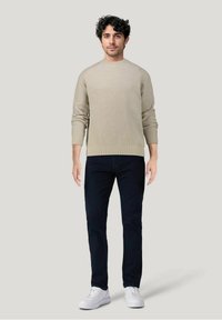 Maglione a coste beige con collo rotondo, abbinato a jeans scuri e sneakers bianche, indossato da un uomo in piedi davanti a uno sfondo neutro.