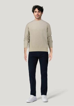 Maglione a coste beige con collo rotondo, abbinato a jeans scuri e sneakers bianche, indossato da un uomo in piedi davanti a uno sfondo neutro.