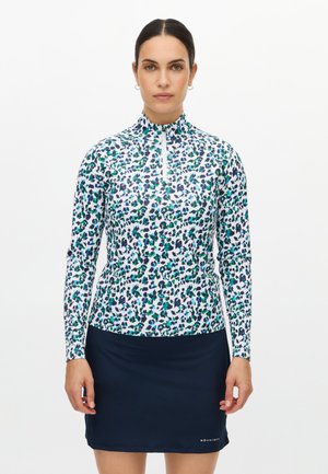 Langärmliges Sportoberteil mit hohem Kragen, weißem Grundton und einem Leopardenmuster in Grün, Blau und Schwarz. Der Rock darunter ist marineblau.