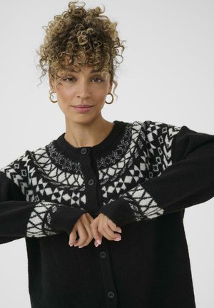 Vrouw met krullend haar die een zwarte cardigan met witte Noorse patronen draagt en de voorkant sluit tegen een effen achtergrond.