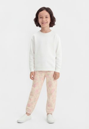 Παιδί με μεσαίου μήκους σκούρα μαλλιά, που φοράει λευκό φούτερ, ροζ φλοράλ παντελόνι και λευκά παπούτσια χωρίς κορδόνια, που χαμογελά και στέκεται σε λευκό φόντο.