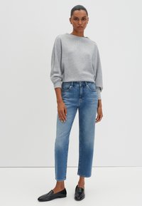 Femme debout, les bras détendus, portant un haut à manches longues gris clair, un jean cropped bleu et des chaussures noires à enfiler sur fond blanc.
