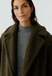 Olivgrüner, strukturierter Mantel mit einemreversierten Kragen über einem hellgrauen, gerippten Rollkragenpullover. Nahaufnahme des Oberkörpers, neutraler Hintergrund.