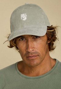 Casquette en velours côtelé verte avec un logo « PULL » brodé sur le devant, visière courbée et tissu texturé. Portée avec une chemise verte assortie.