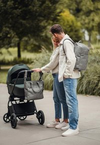 Zelený kočárek s elegantním designem, připevněná šedá textilní taška a černá kola. Vedle něj stojí dva lidé, oba v neformálním oblečení.