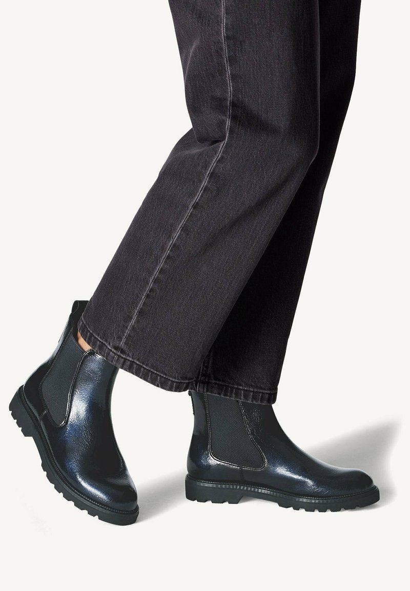 Bottes chevilles en cuir noir à finition brillante, avec des panneaux latéraux élastiques et des semelles robustes, associées à un pantalon court foncé.