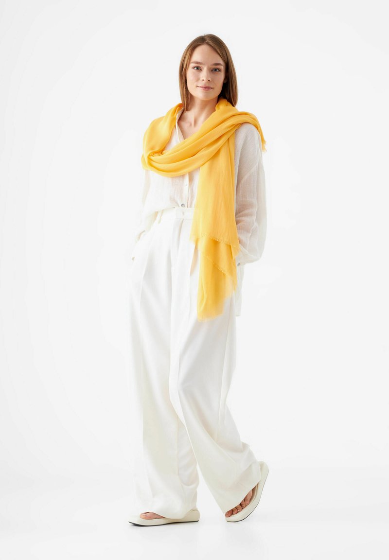 Gele sjaal draped over een witte, langemouwen shirt en hoge taille witte broek, gecombineerd met witte instappers. Soepele textuur, ontspannen pasvorm.