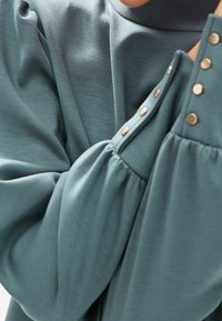 Haut à manches longues couleur sarcelle avec manches froncées, en tissu lisse, doté de boutons décoratifs dorés le long du poignet.