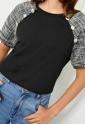 Femme portant un haut noir avec des manches courtes texturées noir et blanc ornées de boutons argentés décoratifs, associée à un jean bleu.