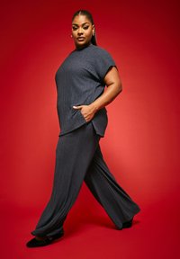 Harmaa teksturoitu yläosa ja leveälahkeiset housut, joissa on lyhyet hihat ja sivutaskut, yhdistettynä mustiin tyyliin, punaisen taustan edessä.