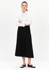 Witte lange mouwen knoopshirt gecombineerd met een zwarte midi-jurk met knopen aan de voorkant en plooien, aangevuld met zwart-witte platte schoenen.