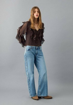 Blusa nera con maniche lunghe arricciate, abbinata a jeans a gamba larga azzurri con strisce laterali e stivaletti marroni alla caviglia.