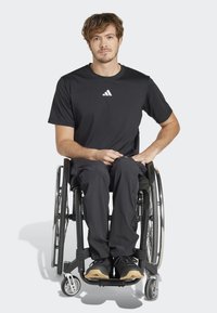 Černé sportovní tričko s bílým logem, spárované s černými kalhotami. Osoba sedí na invalidním vozíku a má na sobě černé tenisky.